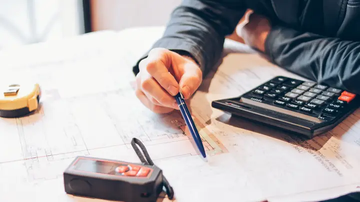 Hand marking a drawing with a pen next to a calculator and laser measure