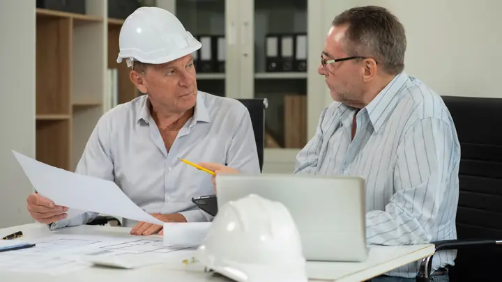 Two older men discussing a document in an office with a white hard hat