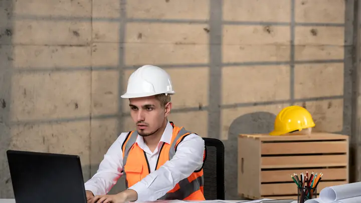Young engineer in a hard hat and orange vest working on a laptop in a site office