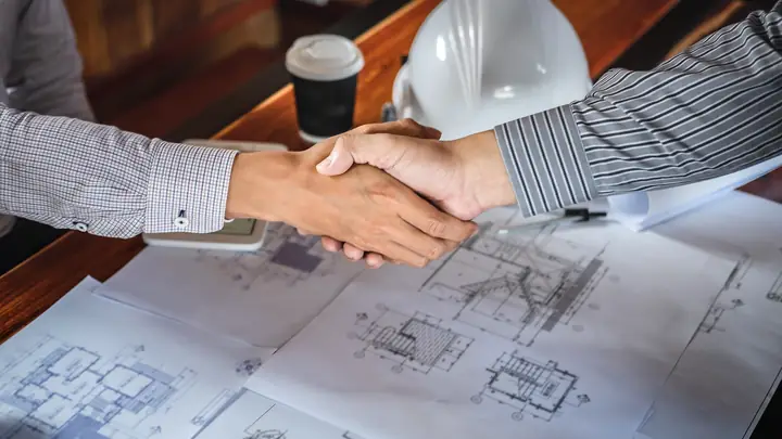 Handshake over detailed drawings with a white hard hat and coffee