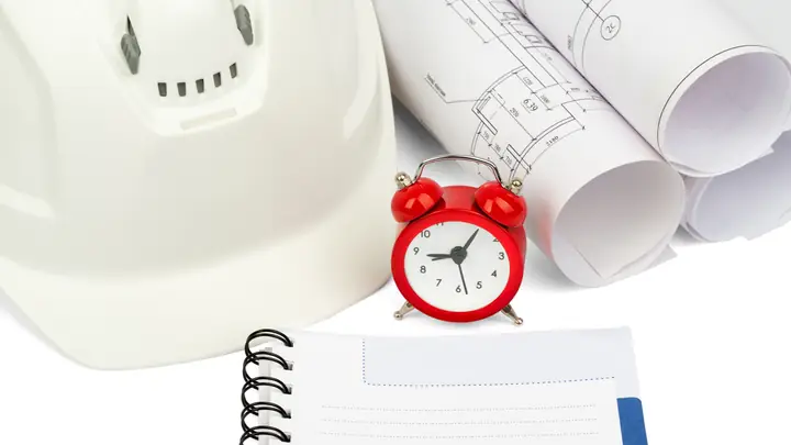 White hard hat, red alarm clock, notebook and rolled drawings
