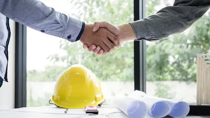 Handshake over a desk with a yellow hard hat and rolled drawings