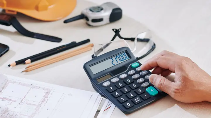 Hand using a calculator beside pencils, safety glasses and an orange hard hat