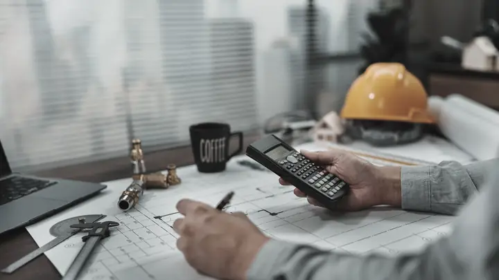 Person using a calculator over drawings with a yellow hard hat on the desk