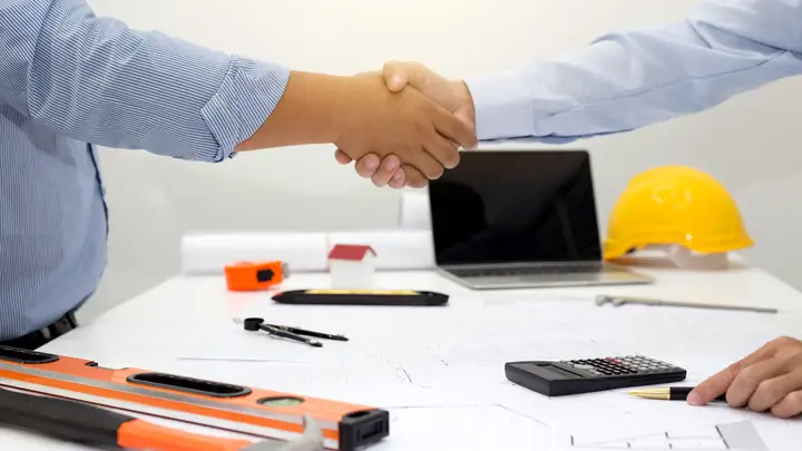 Handshake over a desk with a laptop, level, calculator and hard hat