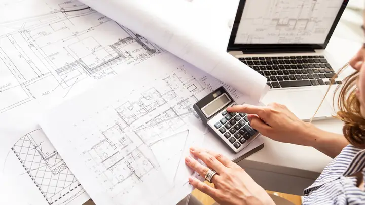 Hands on a calculator over floor plans with a laptop showing a drawing