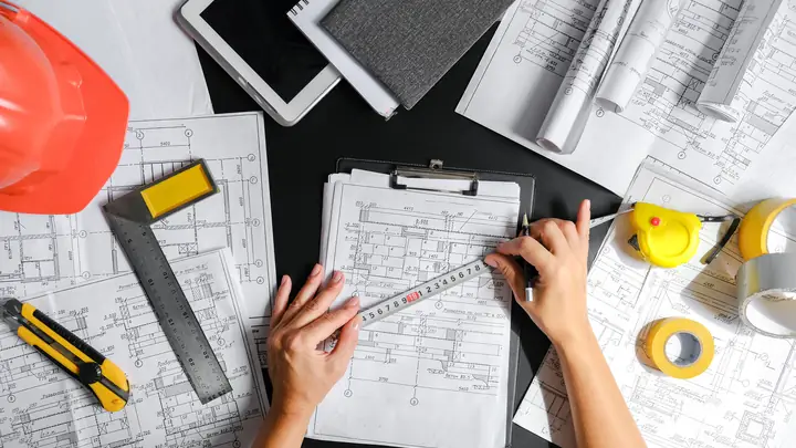 Overhead view of hands measuring a floor plan beside a hard hat, square and tape