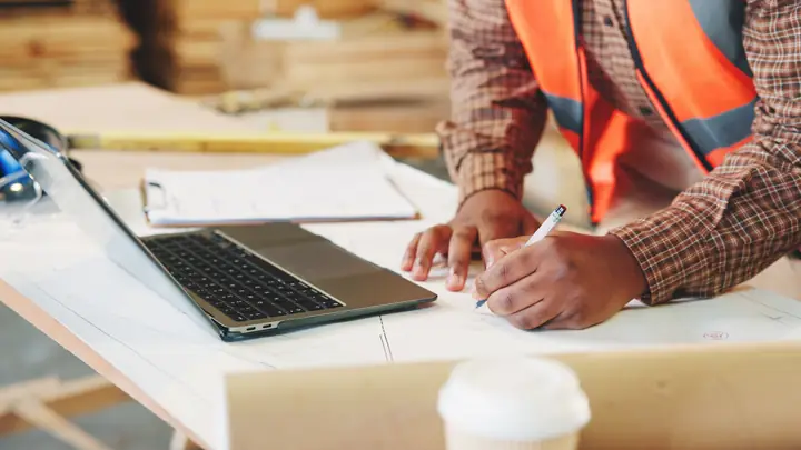 Worker in an orange hi-vis vest writing notes beside a laptop