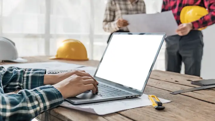 Person at a laptop with a colleague holding drawings and hard hats on the desk