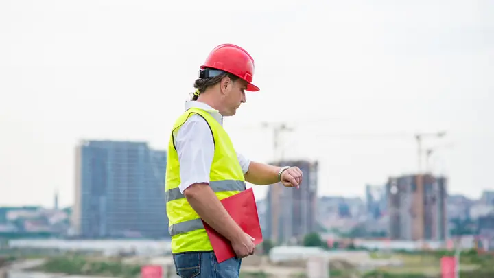 Worker in a red hard hat checking his watch on a site with cranes behind