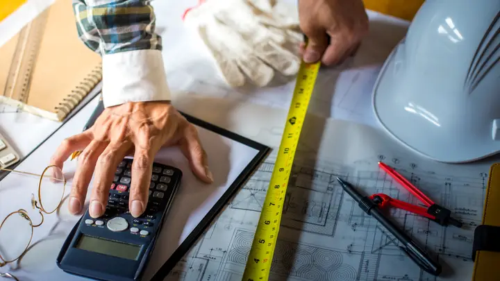 Hand on a calculator with a tape measure, gloves and hard hat on drawings