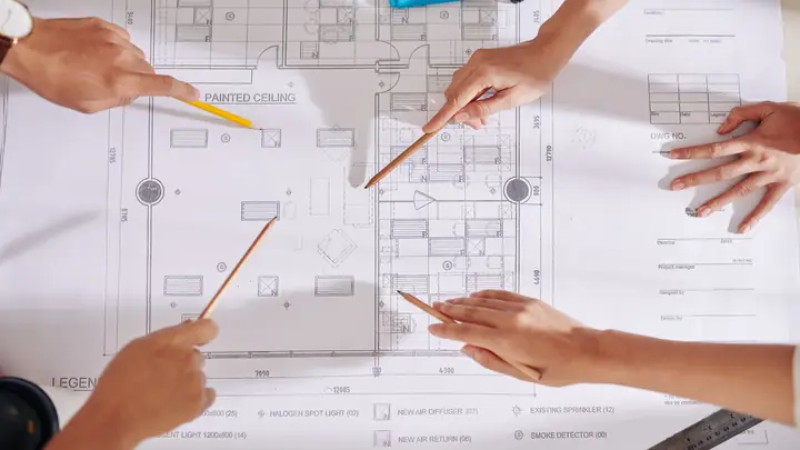 Several hands pointing with pencils at a detailed floor plan