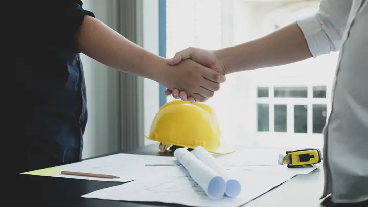 Handshake over a desk with a yellow hard hat and rolled drawings