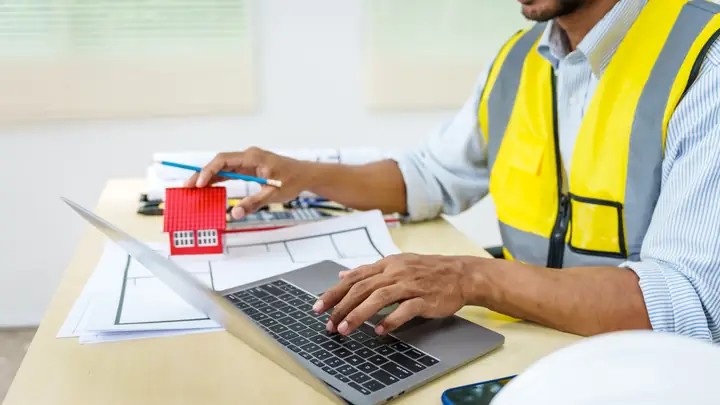 Worker in a hi-vis vest using a laptop at a desk with a small red house model