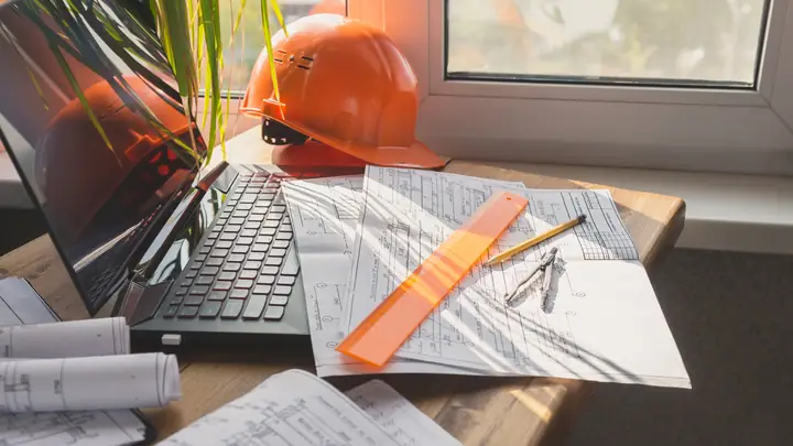 Laptop, orange hard hat and drawings on a desk by a window