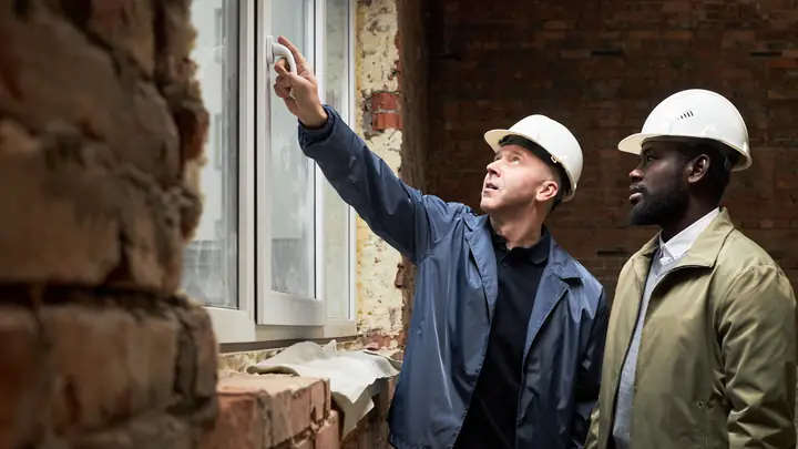 Two men in hard hats looking up at a window in an old brick wall