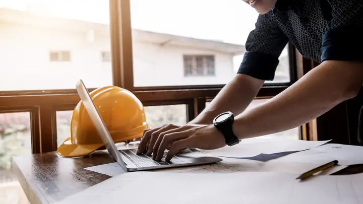 Hands typing on a laptop by a window with a yellow hard hat