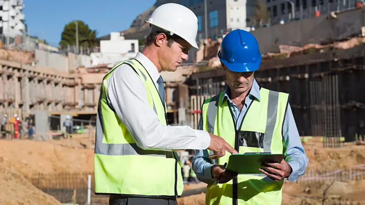 Two engineers in hard hats reviewing a tablet on a city construction site
