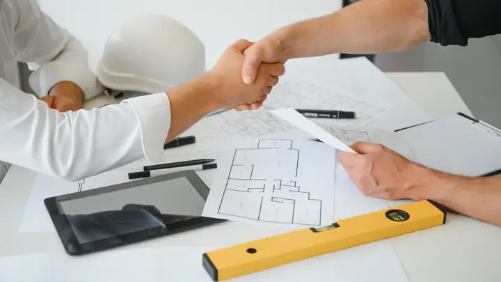 Handshake over a desk with drawings, a tablet and a level