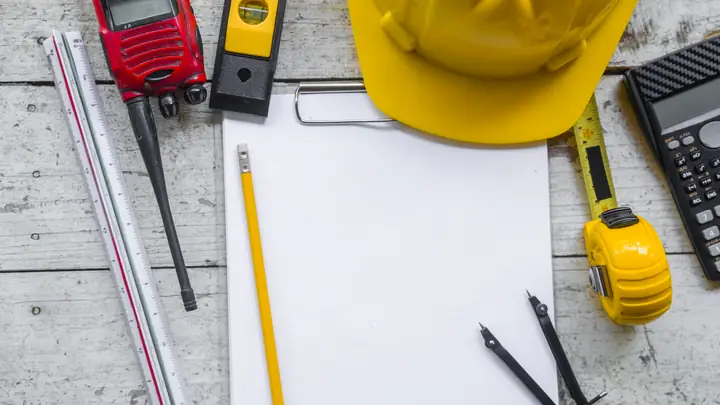 Yellow hard hat, radio, level, calculator and blank paper on a wooden desk