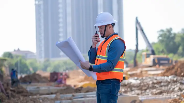 Engineer in a hard hat holding drawings and a radio on a site