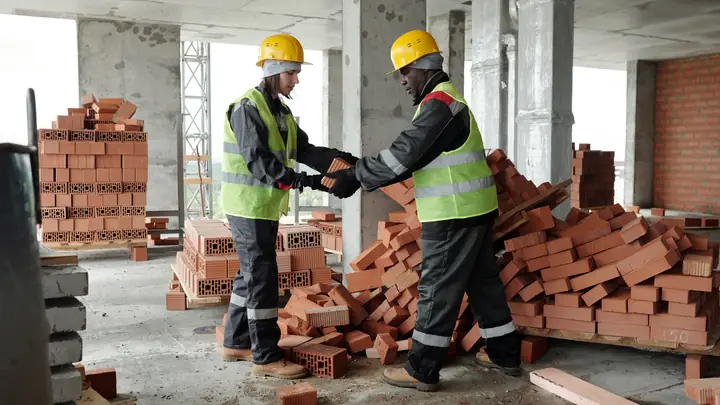 Two workers in hard hats shaking hands among stacks of bricks