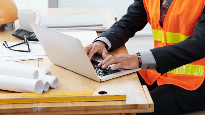 Person in an orange hi-vis vest and suit typing on a laptop at a desk