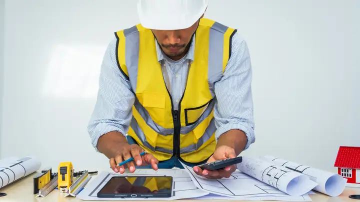 Worker in a hard hat and hi-vis vest using a calculator and tablet at a desk