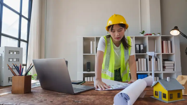 Woman in a hard hat leaning over drawings at a desk with a laptop