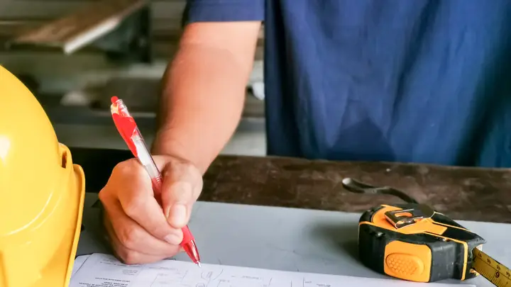 Hand ticking a checklist with a red pen, tape measure and hard hat beside