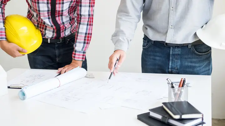Two men at a desk with drawings, one holding a yellow hard hat