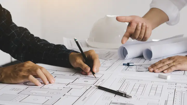 Hands pointing at drawings with a pen, white hard hat behind