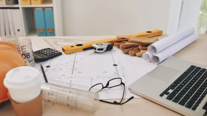 Desk with a laptop, calculator, level, drawings and a coffee cup