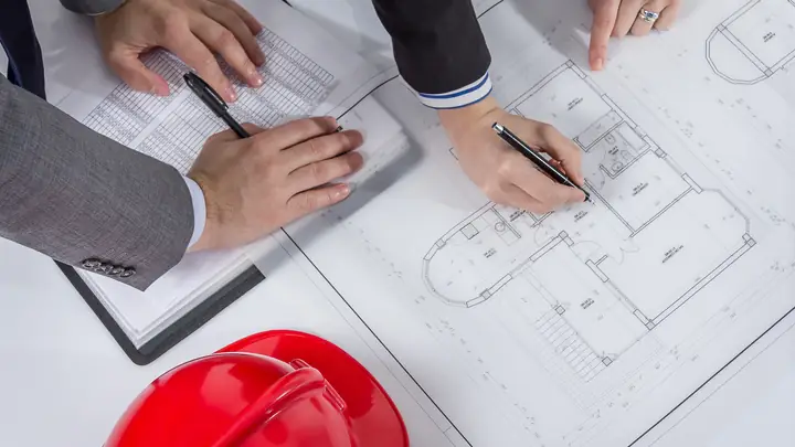 Hands with pens on floor plans beside a red hard hat