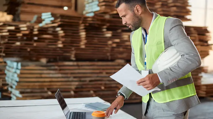 Man in hi-vis with drawings and a laptop in a timber warehouse