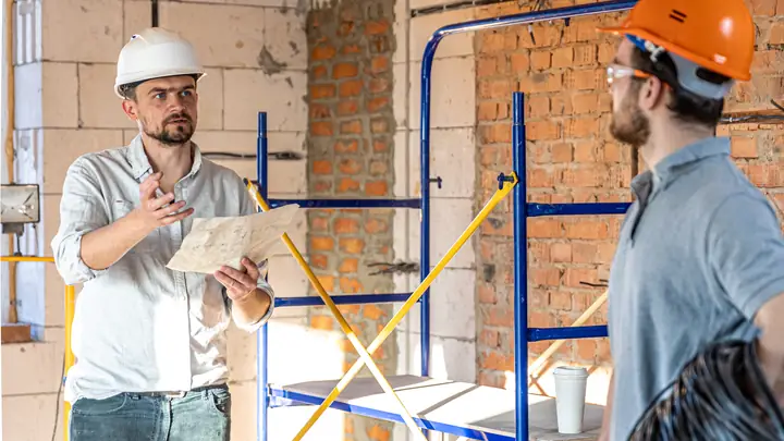 Two tradesmen discussing a drawing by a scaffold tower in a brick interior