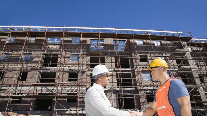Two men in hard hats shaking hands in front of a building wrapped in scaffolding