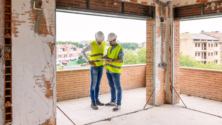 Two men in hi-vis vests inside an unfinished brick room overlooking a town