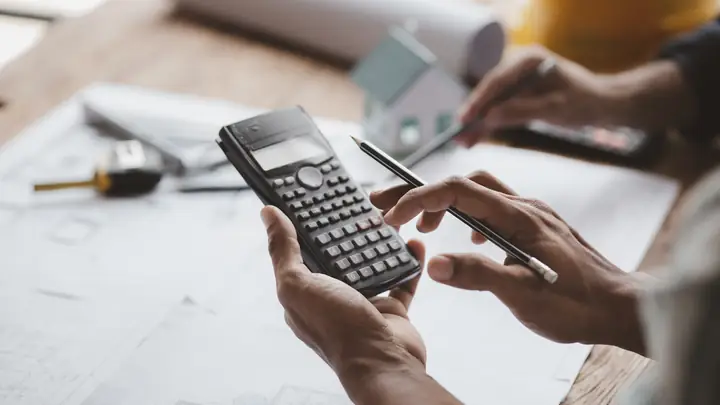 Hands with a calculator and pen over drawings and a small house model