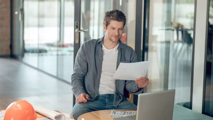 Man reading a document at a laptop with an orange hard hat on the table
