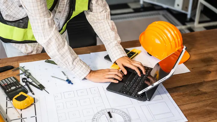 Worker in hi-vis typing on a laptop over drawings with an orange hard hat
