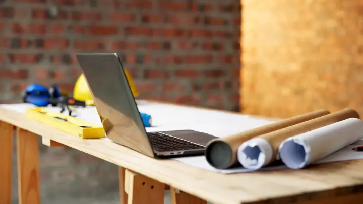 Laptop, rolled drawings and a hard hat on a trestle table against a brick wall