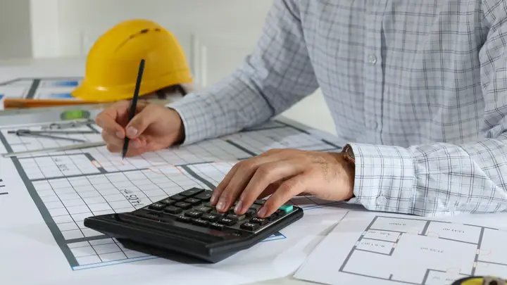 Man using a calculator and pencil over floor plans with a hard hat on the desk