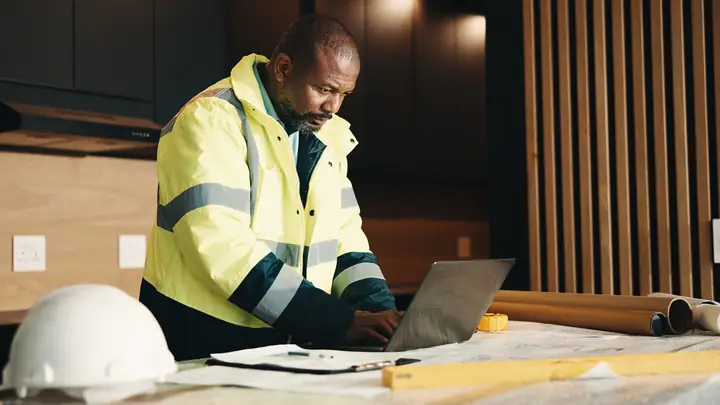 Man in a hi-vis jacket using a laptop at a desk with a white hard hat