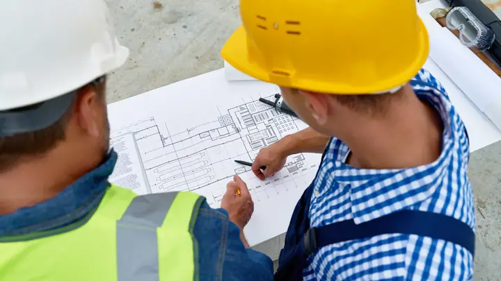 Over-the-shoulder view of two workers in hard hats marking up a drawing
