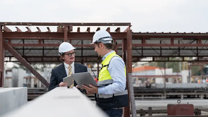 Two men in hard hats with a laptop on a steel-framed site