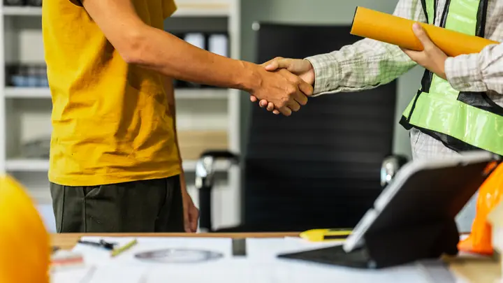 Handshake in an office, one person in a hi-vis vest holding rolled drawings