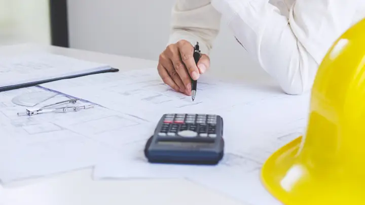 Hand with a pen next to a calculator and yellow hard hat on a set of drawings