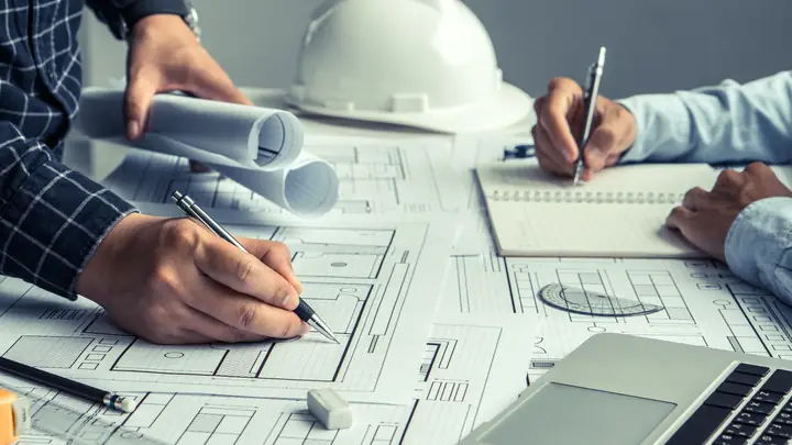 Two people writing on drawings next to a laptop and white hard hat