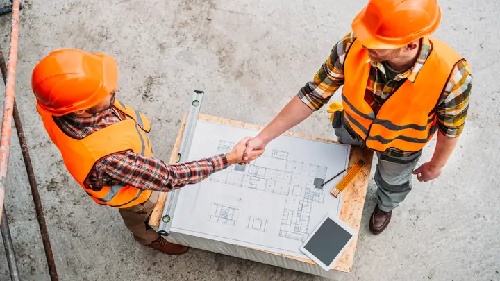 Overhead view of two workers in orange hard hats shaking hands over drawings